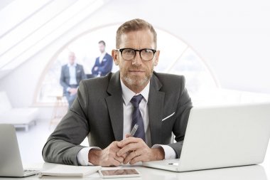 Takım elbise ve dizüstü bilgisayar önünde ofisinde otururken kameraya bakarak başarılı Olgun çift Finans Müdürü iş adamı.