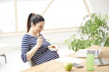 Güzel genç hamile kadın laptop önünde oturan ve evde rahatlatıcı bir tabak meyve salatası yemek. 