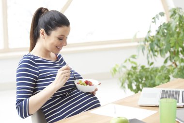 Güzel genç hamile kadın laptop önünde oturan ve evde rahatlatıcı bir tabak meyve salatası yemek. 