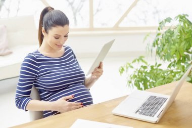 Evde masa başında otururken dijital tablet ve dizüstü bilgisayar kullanan güzel hamile kadın.