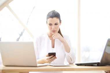 Onun cep telefonu ve dizüstü bilgisayarlar önünde ofis masasında otururken mesajlaşma metin kullanan düşünme genç kadın portresi. Gülümseyen iş kadını evden onun iş yönetme. 