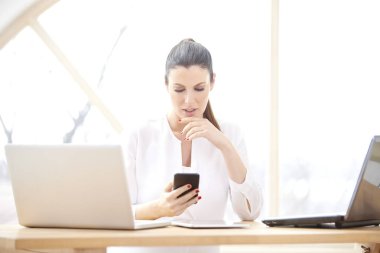 Onun cep telefonu ve dizüstü bilgisayarlar önünde ofis masasında otururken mesajlaşma metin kullanan düşünme genç kadın portresi. Gülümseyen iş kadını evden onun iş yönetme. 