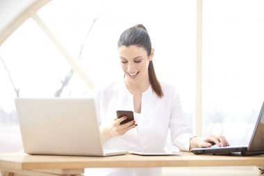 Onun cep telefonu ve dizüstü bilgisayarlar önünde ofis masasında otururken mesajlaşma metin kullanan güzel genç kadın portresi. Gülümseyen iş kadını evden onun iş yönetme. 