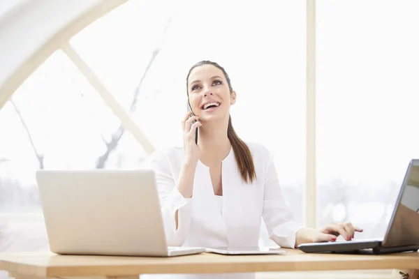 快乐的年轻女商人 在办公室里坐在笔记本电脑前 用手机和客户咨询 — 图库照片