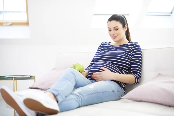 快乐怀孕的妇女手拿着一个苹果 而坐在沙发上放松 — 图库照片