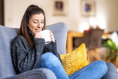 Evdeki koltukta dinlenirken ve çay içerken boğazlı kazak ve kot pantolon giyen gülümseyen kadın..