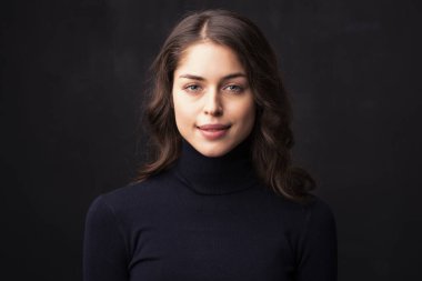 Güzel gülüşlü, balıkçı yaka kazak giyen, koyu arkaplanda poz veren çekici genç bir kadının portresi.. 