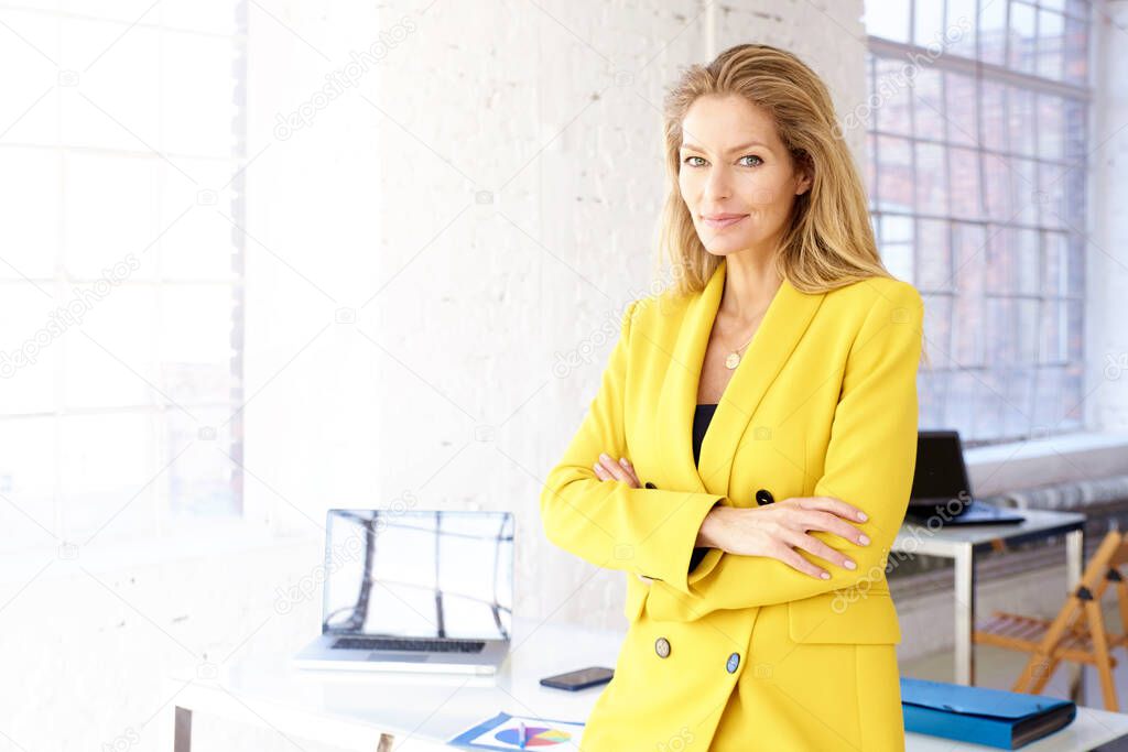 Portrait shot of attractive mature businesswoman wearing yellow blazer while standing at office desk. 