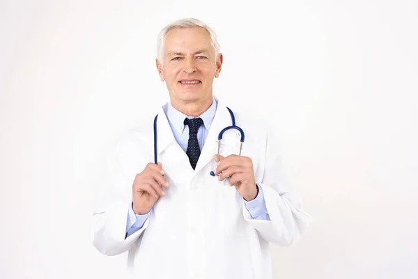 白い背景に立っている間彼の首の周りに制服を身に着けている彼の首の周りに聴診器でシニア男性医師の肖像写真 — ストック写真