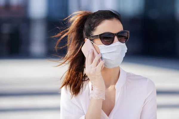Close-up shot of confident woman wearing face mask and talking with somebody on her mobile phone while walking in front of business center during coronavirus pandemic.