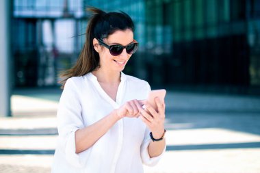 Sokakta durup mesajlaşırken güneş gözlüğü ve beyaz gömlek takan mutlu bir kadının fotoğrafı..