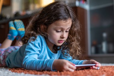 Evdeki oturma odasında dijital akıllı telefon kullanarak uzanan sevimli küçük mutlu kız. Aile etkinliği kavramı.