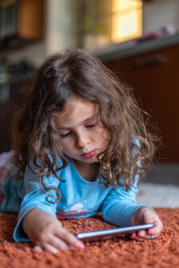Evdeki oturma odasında dijital akıllı telefon kullanarak uzanan sevimli küçük mutlu kız. Aile etkinliği kavramı.