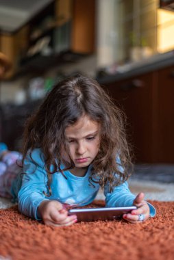 Evdeki oturma odasında dijital akıllı telefon kullanarak uzanan sevimli küçük mutlu kız. Aile etkinliği kavramı.