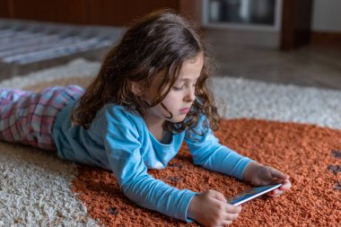 Evdeki oturma odasında dijital akıllı telefon kullanarak uzanan sevimli küçük mutlu kız. Aile etkinliği kavramı.