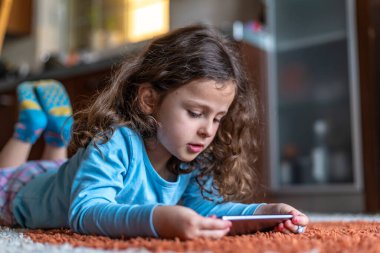 Evdeki oturma odasında dijital akıllı telefon kullanarak uzanan sevimli küçük mutlu kız. Aile etkinliği kavramı.