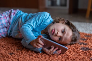 Evdeki oturma odasında dijital akıllı telefon kullanarak uzanan sevimli küçük mutlu kız. Aile etkinliği kavramı.