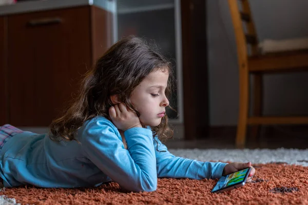 Evdeki oturma odasında dijital akıllı telefon kullanarak uzanan sevimli küçük mutlu kız. Aile etkinliği kavramı.
