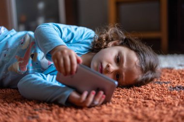 Evdeki oturma odasında dijital akıllı telefon kullanarak uzanan sevimli küçük mutlu kız. Aile etkinliği kavramı.