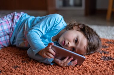 Evdeki oturma odasında dijital akıllı telefon kullanarak uzanan sevimli küçük mutlu kız. Aile etkinliği kavramı.