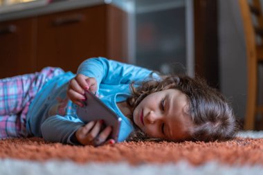 Evdeki oturma odasında dijital akıllı telefon kullanarak uzanan sevimli küçük mutlu kız. Aile etkinliği kavramı.