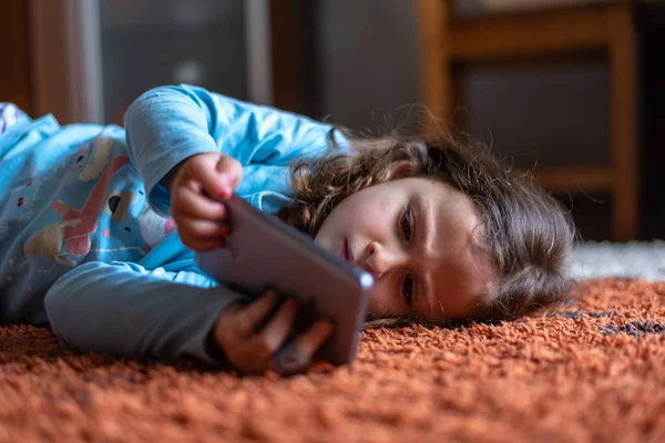 Evdeki oturma odasında dijital akıllı telefon kullanarak uzanan sevimli küçük mutlu kız. Aile etkinliği kavramı.