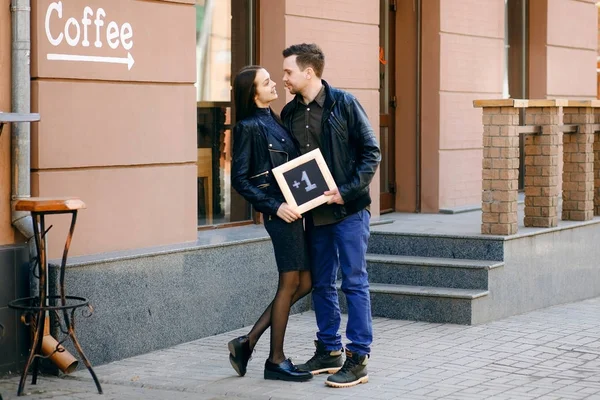 Ένα ευτυχισμένο παντρεμένο ζευγάρι που στέκεται στο δρόμο και κρατώντας μια μαύρη πλάκα με επιγραφή συν ένα — Φωτογραφία Αρχείου