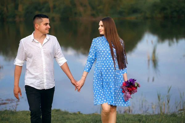 Egy fiú és egy lány sétálnak a folyóparton, fogják egymás kezét, egy lány virágokkal, Valentin-nap. — Stock Fotó