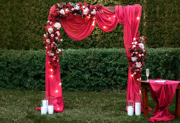 Свадьба. Церемония. Свадебная арка. Красная свадебная арка цветов и зелени стоит на зеленой траве в парке — стоковое фото