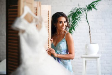 Güzel bir kız düğüne hazırlanıyor, askıda asılı bir elbise, beyaz bir duvara poz veren esmer bir kız.