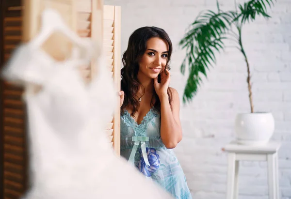 Menina bonita se preparando para o casamento, um vestido pendurado em um cabide, uma morena posando contra uma parede branca — Fotografia de Stock