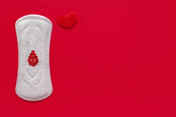 Menstrual pad with red sequins in the shape of a heart on a red background, top view, miniature of the menstrual period — Stock Fotó