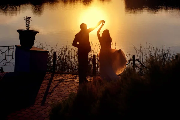Silhouettes of the bride and groom, man and woman dancing by the river at sunset, beautiful bride and courageous groom, summer sun, dance of love
