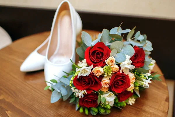 新娘的白鞋和一束红玫瑰躺在宾馆房间的桌子上 新娘的早晨 婚礼的准备工作 婚礼的附件 新娘的花束 — 图库照片