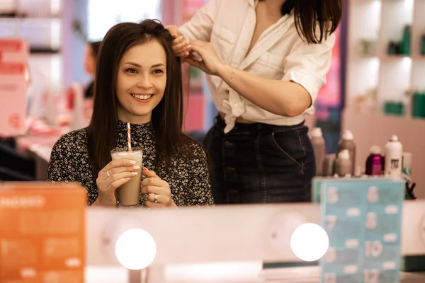 美容室でブルネットにヘアスタイルをする女性の美容師 ヘアスタイルを作成するプロセス プロの美容師は髪のボリュームを作成し クライアントの女の子は笑顔とコーヒーを飲みます — ストック写真