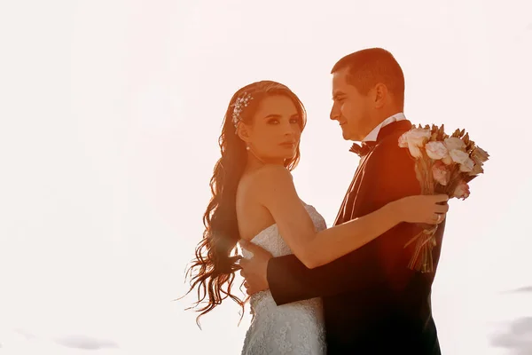 The bride and groom are hugging in the desert against the sunset, a couple in love, a beautiful wedding dress, orange light on their faces, the wedding couple is enjoying each other, a wedding bouquet