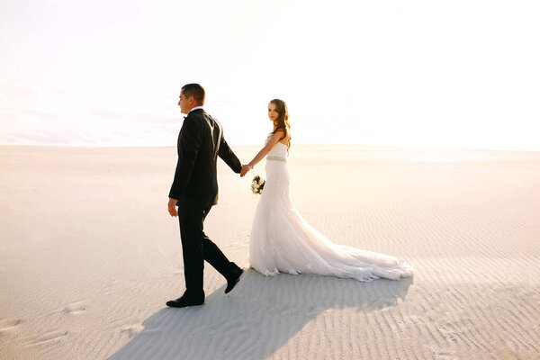 The bride and groom are walking in the desert, blue sky and white sand, a beautiful couple, wedding day, pursuit of goals, survival, thirst, low clouds, desert dunes, beautiful sunset