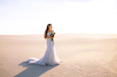 Uzun bir gelinlik ve bir buket ile gelin çöldeki kumda duruyor, gelinin güzel bir buketi, tasarımcı gelinliği, özgürlük hissi, hassas bir kız eş oluyor.