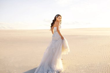 Güzel, uzun elbiseli bir gelin çölde kumların üzerinde yürüyor, arka plan, gün batımı, hassas kız, tasarımcı gelinliği, özgürlük hissi, rüzgar saçlarını hareket ettiriyor, çöl kumları...