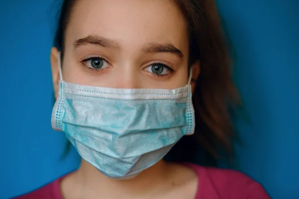 Girl in a protective medical mask on a blue background, protection against airborne infection, global epidemic of coronovirus, pandemic covid -19, home quarantine, sad eyes, close-up