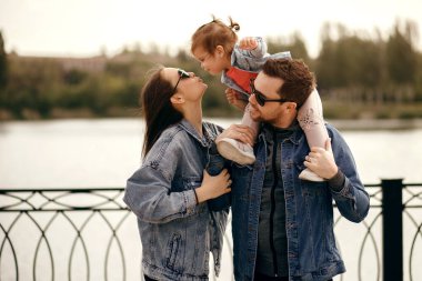 Küçük kız babasının omuzlarında oturur ve annesini öper, şık ebeveynler kot pantolon giyer, güneş gözlüğü takar, güzel genç bir aile nehir kenarında yürür ve günbatımında eğlenir.