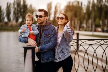 Aile yürüyüşü, baba ve anne kızı kollarında tutuyor, gün batımı, kot elbiseli aile, güneş gözlüğü, sonbahar havası, şık bir aile, güzel bir çift, anne saçını düzleştiriyor.
