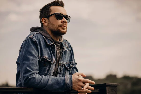 Portrait of a stylish handsome man with stubble. A man wears jeans clothes and sunglasses. A guy stands on the embankment of the river and looks into the distance, copy space