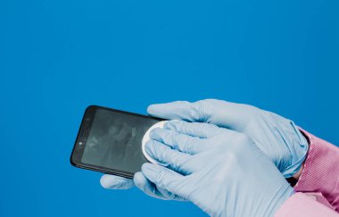 Tıbbi eldivenli bir adam telefonunu pamuk pedlerle siler. Mavi arka plan. Antibakteriyel etki, dezenfekte, dezenfekte