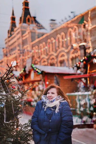 Smiling woman in winter clothes and a headscarf in the Russian style building background with glowing lights. The impression of winter and celebration.