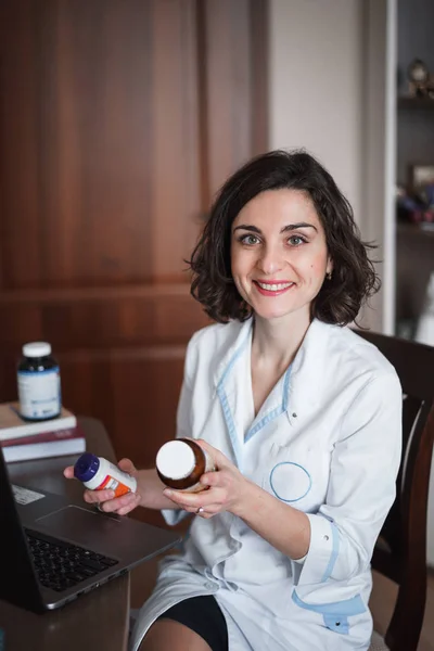 A young smiling brunette doctor woman in white medical gown sits at her desk and holds jars of medicines or vitamins in her hands. Vertical photo.