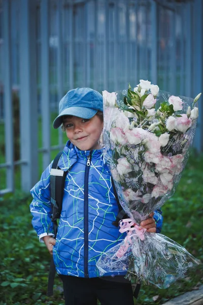 Smiling elementary school pupil with a backpack behind him and a bouquet of flowers in a blue jacket and a cap is standing on the street in front of the school.