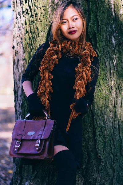 Menina bonita em roupa preta com luvas e bolsa de couro, de pé ao lado da árvore — Fotografia de Stock