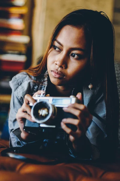 Gorgeous young woman lying on leather sofa holding old Vintage retro film camera in her hands. Fashionable woman Retro style — Stock Photo, Image