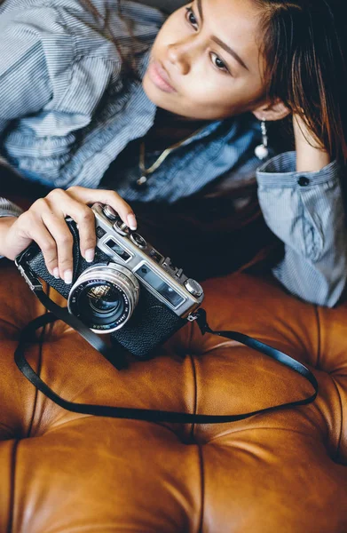 Gorgeous young woman lying on leather sofa holding old Vintage retro film camera in her hands. Fashionable woman Retro style — Stock Photo, Image